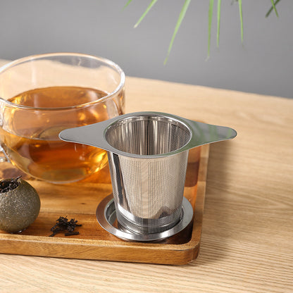 Tea infuser on a wooden tray with a glass of tea and tea leaves in the background.