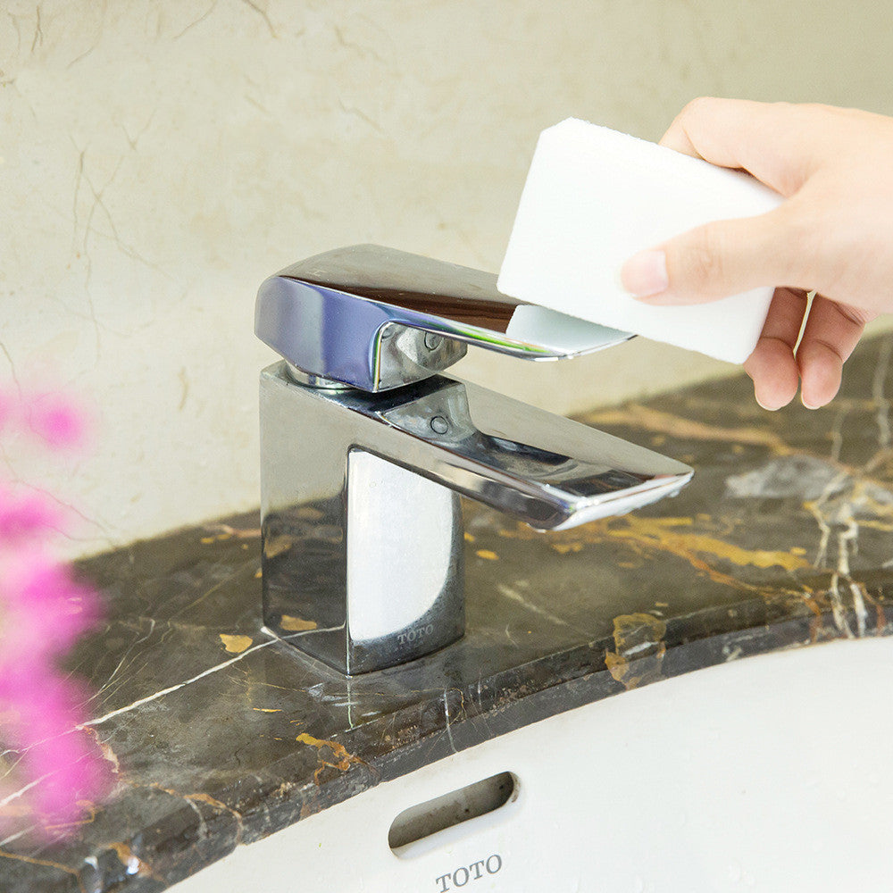 Hand using a white bottle to dispense soap onto a silver faucet on a marble countertop.