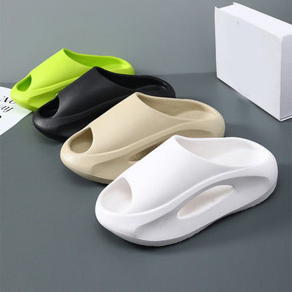 Four slippers in green, black, beige, and white on a gray surface with a white box in the background.